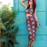fashion portrait of attractive stylish woman posing in summer outfit printed dress, wearing trendy accessories, sandals, purse, sunglasses, red hat, relaxing on vacation
